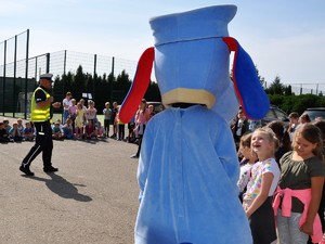 policjant prowadzi spotkanie profilaktyczne z dziećmi, na boisku szkolnym dzieci stoją przed policjantem, obok dzieci stoi maskotka śląskiej Policji, pies Sznupek