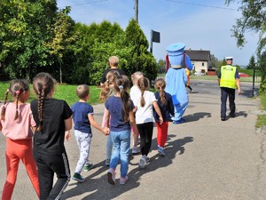 policjant oraz maskotka śląskiej policji wychodzą ze szkolnego boiska, kierują się w stronę przejścia dla pieszych, za nimi idą w parach dzieci