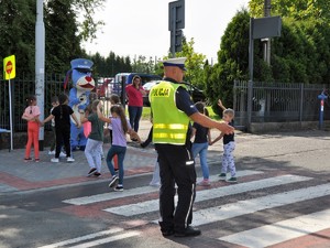 policjant stoi na przejściu dla pieszych, przez przejście dla pieszych przechodzą dzieci, trzymają w górze rękę