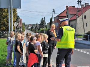 policjant stoi przy przejściu dla pieszych z grupą dzieci, tłumaczy im zasady bezpiecznego poruszania się po drodze